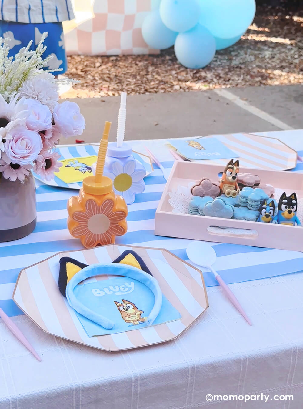 An adorable kid's picnic party table setting with Bluey themed various tablewares from Momo Party including peach and summer blue striped dinner plates, light blue striped table runner, daisy shaped tumbler cups in peach and white, Bluey small napkins, Bluey decorative headbands. Along with a Bluey-inspired daisy shaped macarons, floral bouquet decorations, it's a perfect table setting idea for a modern Bluey themed birthday celebration.