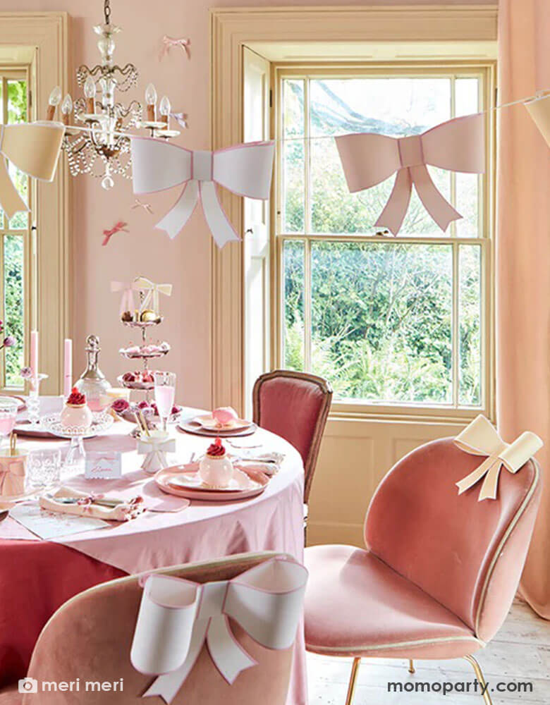 An elegant and chic bow themed party set up featuring Momo Party's pastel heart shaped plates and napkins and 3D bow party cups on the table, above the table there's an elegant chandelier with Meri Meri's 3D bow garland hung over. On the table there are scrumptious treats and sweets for tea time. The chairs are also adorn with 3D bow decoration for a playful touch.