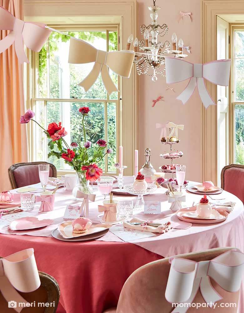 An elegant and chic bow themed party set up featuring Momo Party's pastel heart shaped plates and napkins and 3D bow party cups on the table, above the table there's an elegant chandelier with Meri Meri's 3D bow garland hung over. On the table there are scrumptious treats and sweets for tea time. The chairs are also adorn with 3D bow decoration for a playful touch.