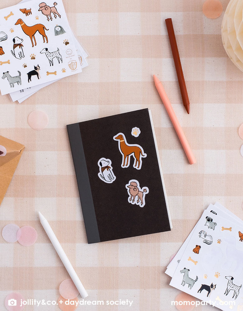 A soft peach checkered table with a small black notebook with Momo Party's Bow Wow dog stickers on it. Around the notebook there are nude/natural colored pencils, giving some inspirations for kid's puppy/dog themed birthday party goodie bag filler ideas.