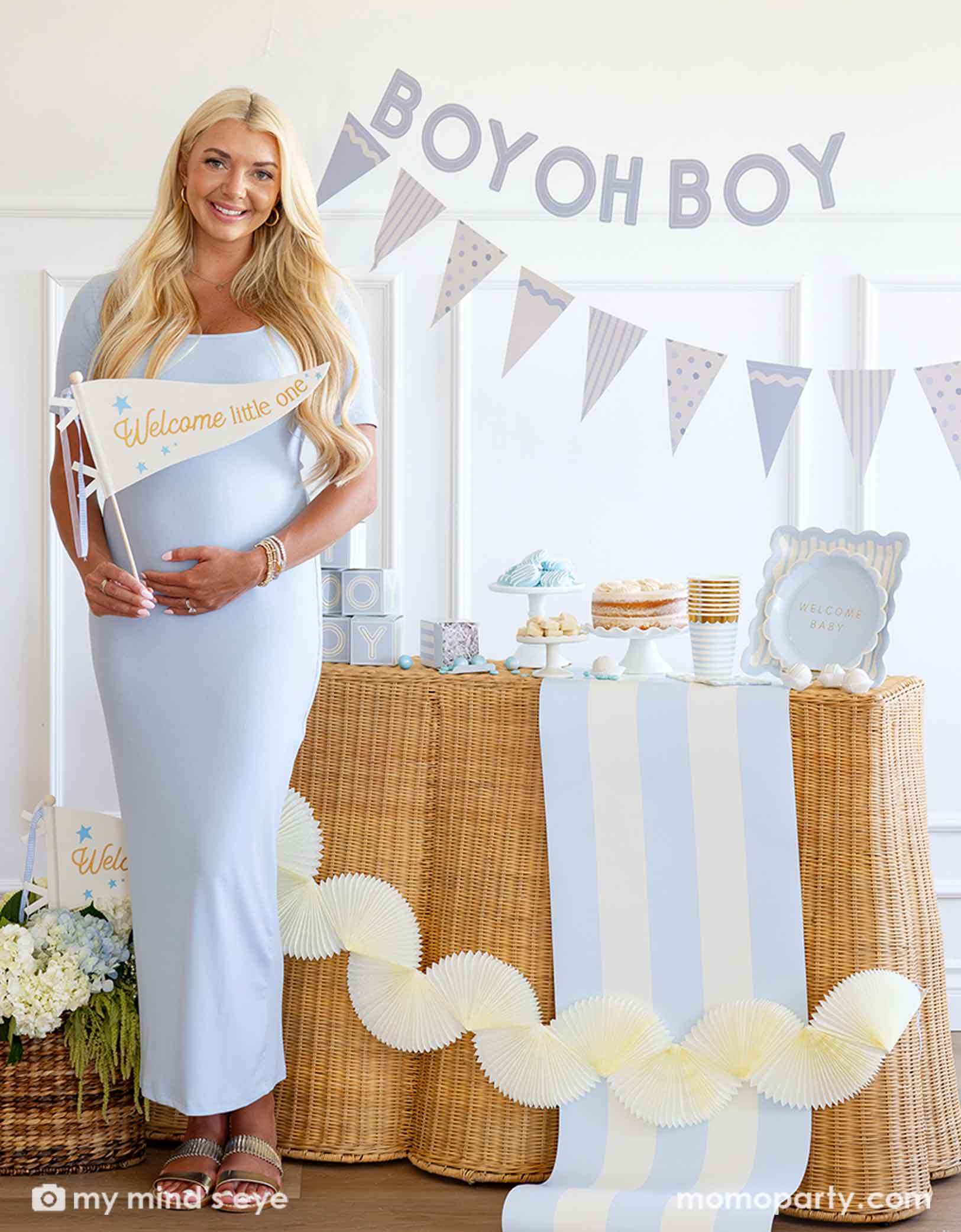 A modern and clean boy baby shower set up featuring various baby blue themed party supplies from Momo Party including party plates, cups, napkins, centerpieces on the table. A pregnant mom-to-be is standing in front of a baby shower setup with 'BOY OH BOY' banner and dessert table. She's holding a party pennant that says "Welcome to the World". With beautiful flower bouquet and cream colored bunting party banner around her.