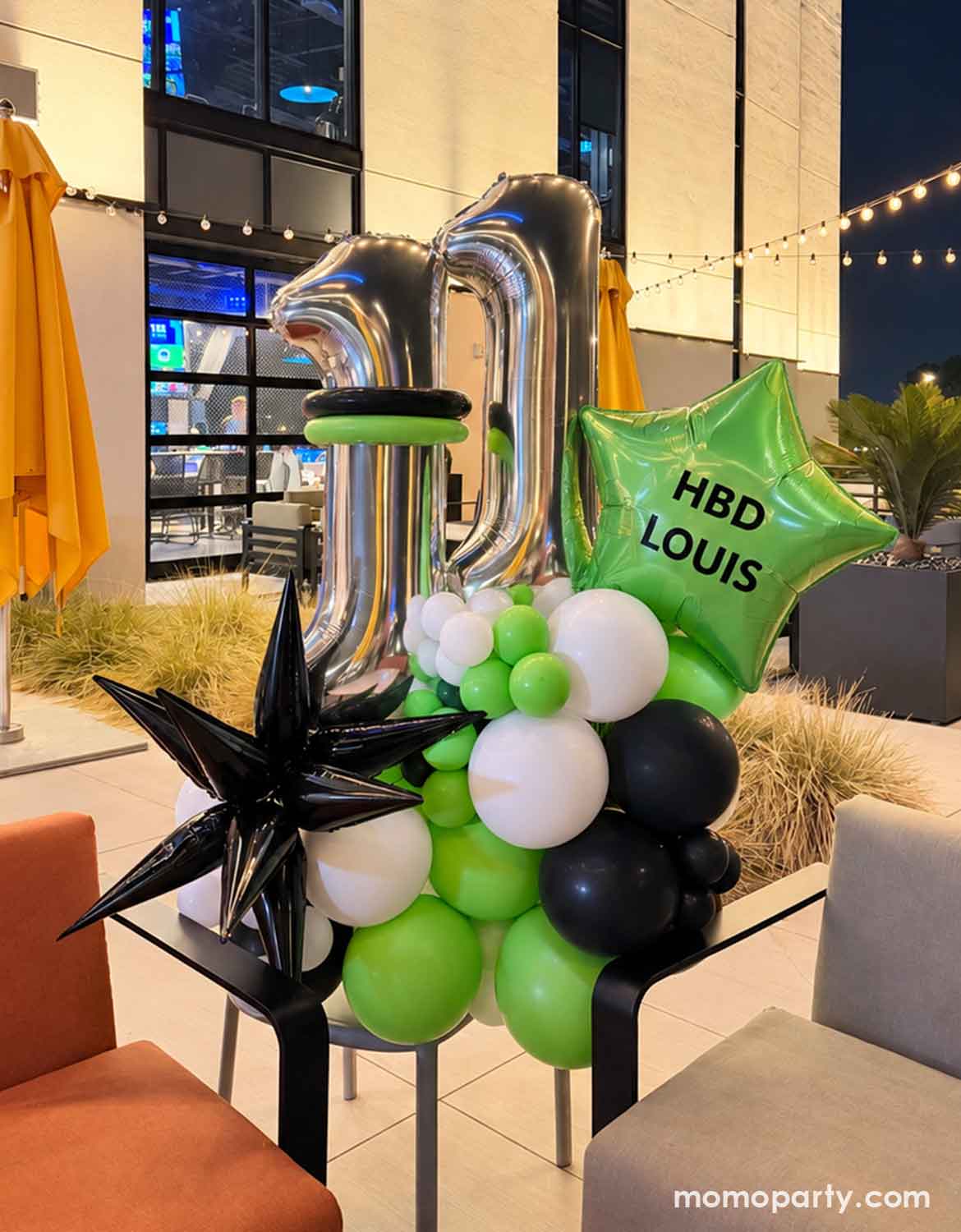 An outdoor party balloon set up featuring a modern and stylish birthday balloon arrangement by Momo Party for a 11th birthday featuring two large metallic silver number 11 foil balloons, a bright green star-shaped balloon with happy birthday wording a black starburst foil balloon decoration, and clusters of black, white, and lime green balloons in a outdoor sitting area next to a lawn.