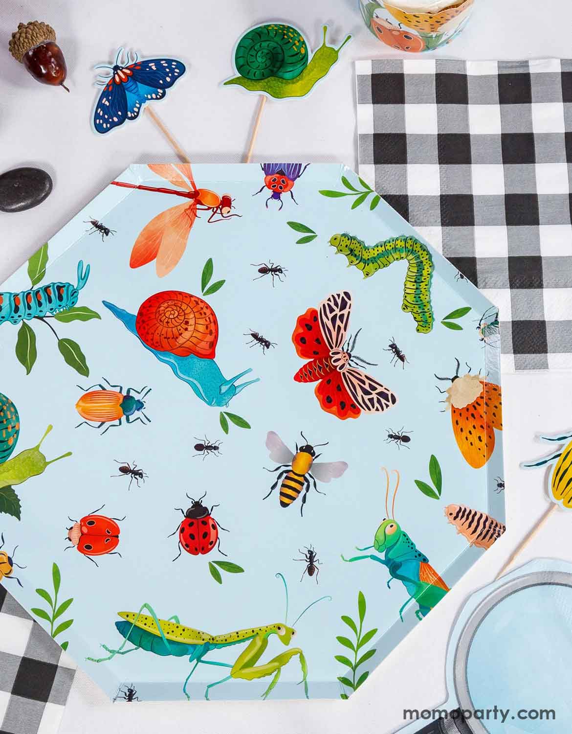 Momo Party's 10" light blue hexagonal party plate with colorful insect pattern next to a checkered paper napkin and insect themed cupcake toppers.