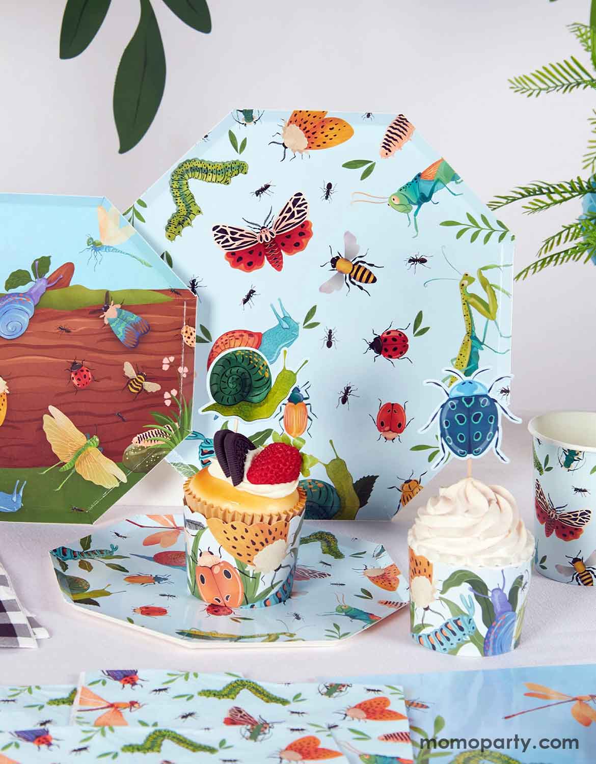 A children's bug themed party table setting with insect-themed tablewares and decorations with natural design elements from Momo Party, including paper plates, cups, napkins, cupcake toppers and wrappers.