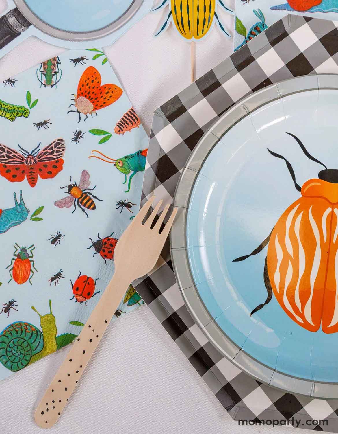 An overhead shot of a children's bug themed party table setting with bug themed party supplies from Momo Party including one beetle round paper plate on checkered hexagonal dinner plate, napkins with insect pattern, magnifying glass shaped napkins, and a wooden fork with black speckle on it on a white table.