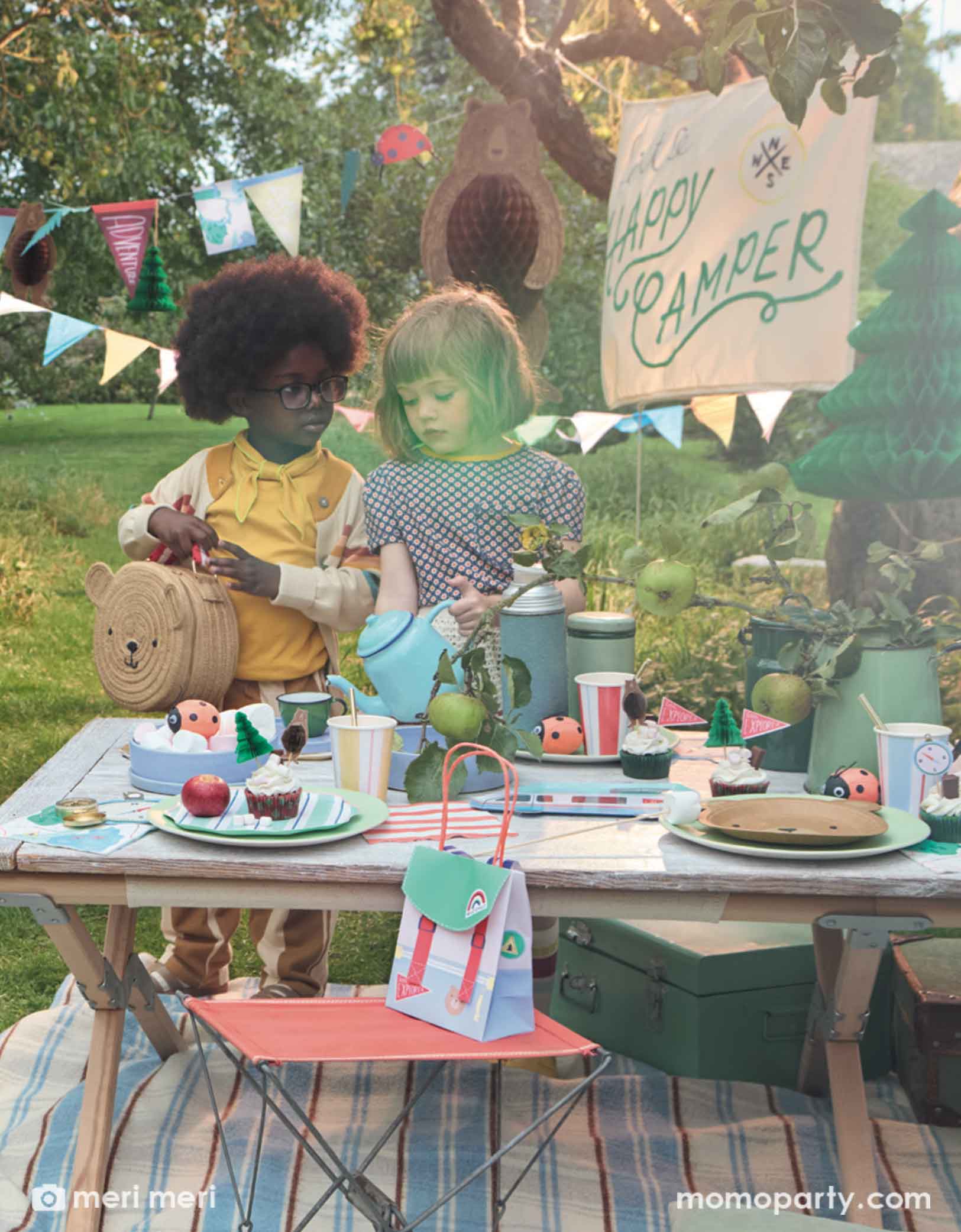 Two cute school aged girls standing next to a camping themed party table featuring multiple Meri Meri camping themed party supplies from Momo Party, including camping RV shaped plates, bear plates, compass shaped napkins, backpack shaped party bags and ladybug surprise balls. Above them hung a 10ft camping icon party garland featuring camping themed pennants and bear & tree honeycomb hanging decorations. With a sign says "Happy Camper" this makes a great inspo for kid's camping themed party decorations. 