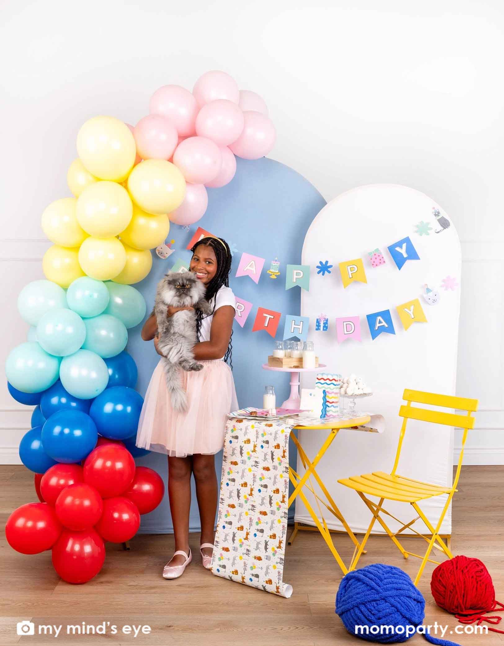 A girl holding a flurry cat in front of a colorful balloon arch with Momo Party's  Cat 'Happy Birthday' party banner hung on the blue and white party backdrop boards. Next to it is a set of yellow table and chair filled with cat themed party supplies from Momo Party including plates, napkins, table runner, and party cups. Along with the jumbo yarns in blue and red on the ground, this sets a great idea for cat themed parties.