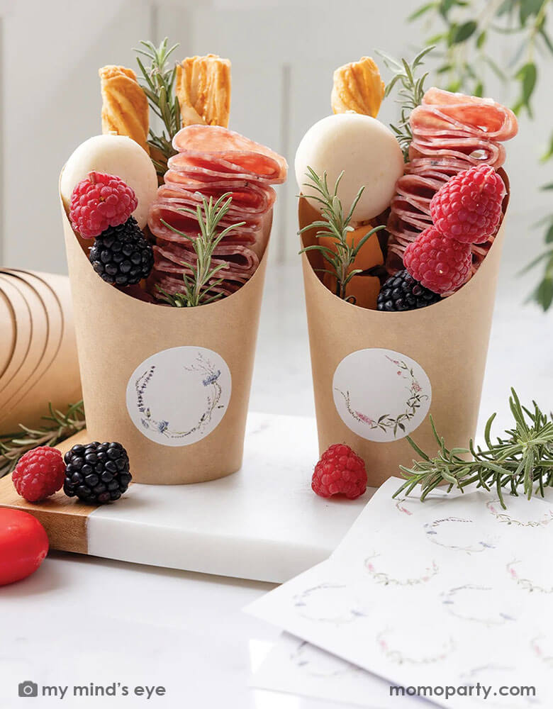 Two charcuterie cups filled with cheese, fruits and salami treats on a white marble kitchen counter. Around them are a marble cutting board with some berries on it and rosemary bunches. These charcuterie cups from Momo Party that come with floral round labels for your guests names are perfect for a spring or summer soriee, whether it's a baby shower, a bridal shower or kid's birthday parties.