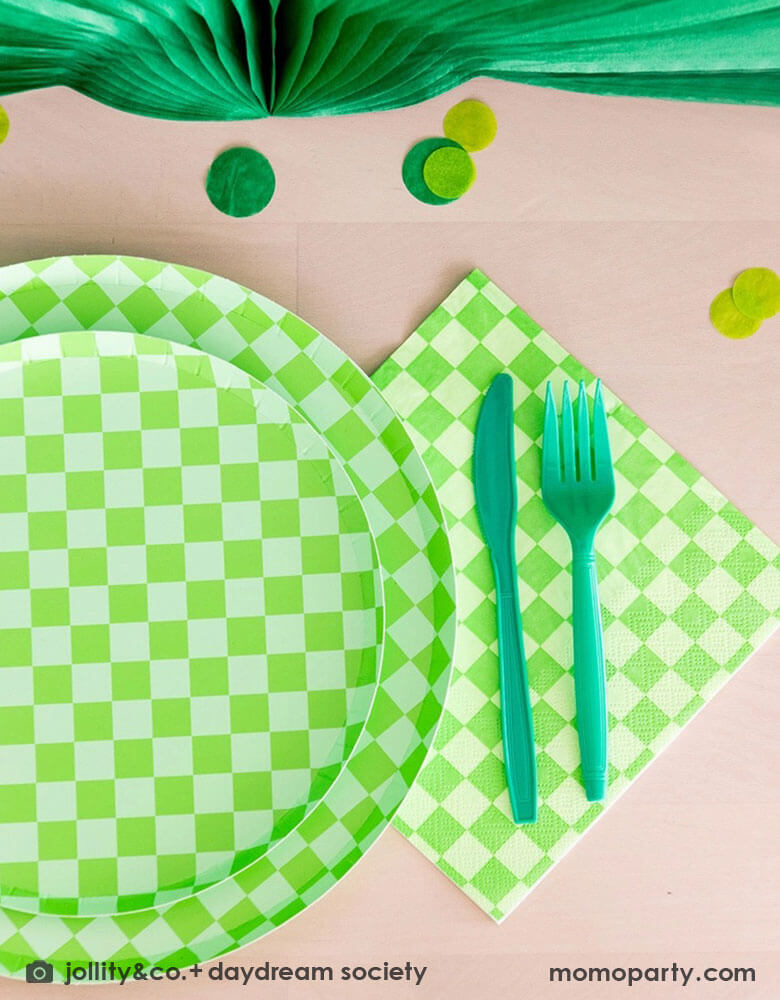 A green themed party table features Momo Party's Check it! Lime Green Checkered Tableware Collection by Jollity & Co. including 10" dinner plates, 8" dessert plates, 6.5" large napkins, green utensils and solid green party cups, with green paper fans as the centerpiece and large round green confetti scattered on the table.