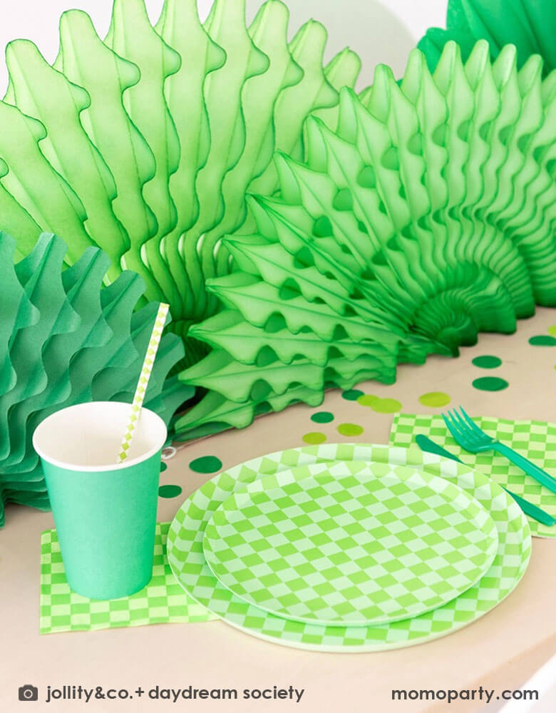 A green themed party table features Momo Party's Check it! Lime Green Checkered Tableware Collection by Jollity & Co. including 10" dinner plates, 8" dessert plates, 6.5" large napkins, green utensils and solid green party cups, with green paper fans as the centerpiece and large round green confetti scattered on the table.