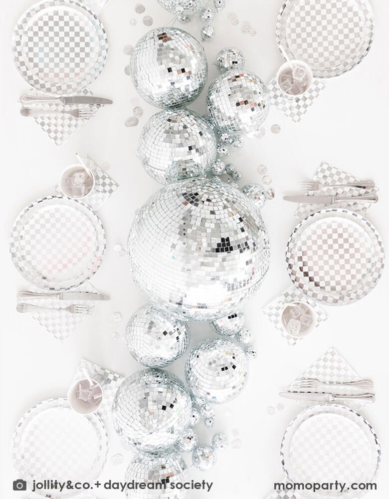 A glamorous and dazzling party table featuring Momo Party's check it! silver checkered tableware collection by Jollity & Co. including 10" dinner plates, 8" dessert plates, 6.5" large napkins, solid silver party cups and silver cutlery. In the middle of the table are a bunch of disco balls in different sizes with silver confetti scattered, what a festive party table! A perfect inspo for New Year's Eve celebration or a disco themed party!