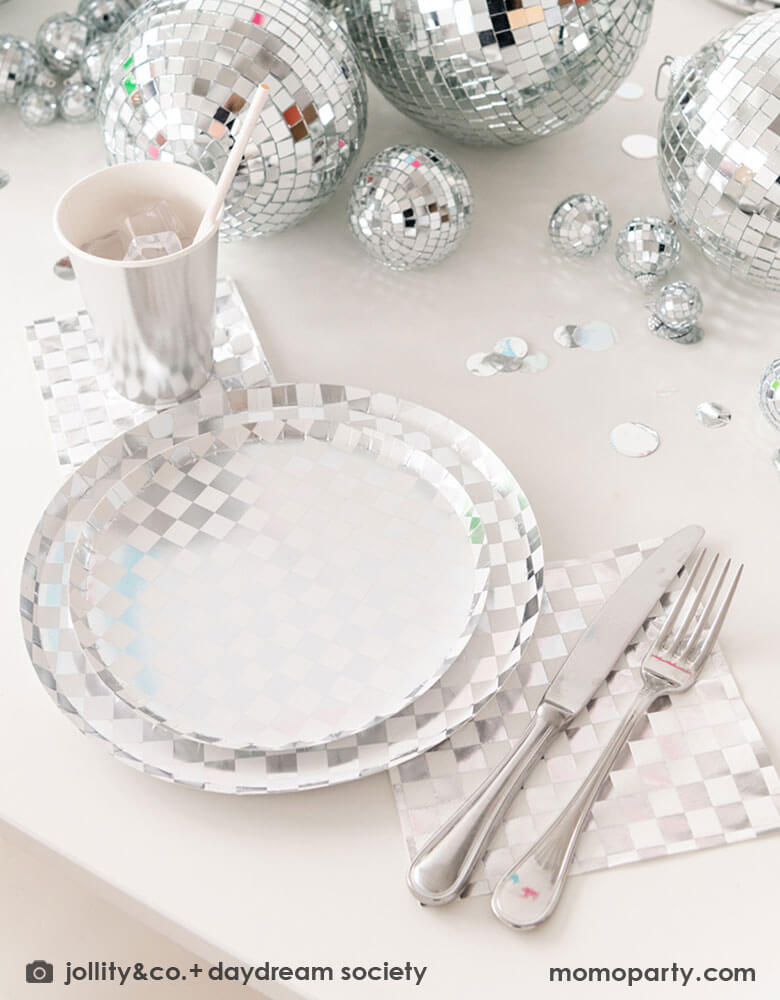 A glamorous and dazzling party table featuring Momo Party's check it! silver checkered tableware collection by Jollity & Co. including 10" dinner plates, 8" dessert plates, 6.5" large napkins, solid silver party cups and silver cutlery. In the middle of the table are a bunch of disco balls in different sizes with silver confetti scattered, what a festive party table! A perfect inspo for New Year's Eve celebration or a disco themed party!