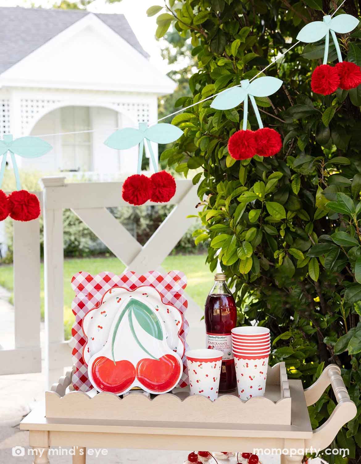 An outdoor party setting next to a tree with cherry-themed decor and party tableware from Momo Party including cherry themed plates, cups, and a bottle of cherry juice on a wooden serving cart. Above it hung a pom pom cherry felt party banner - a simply yet adorable idea for am outdoor cherry themed party in summer.