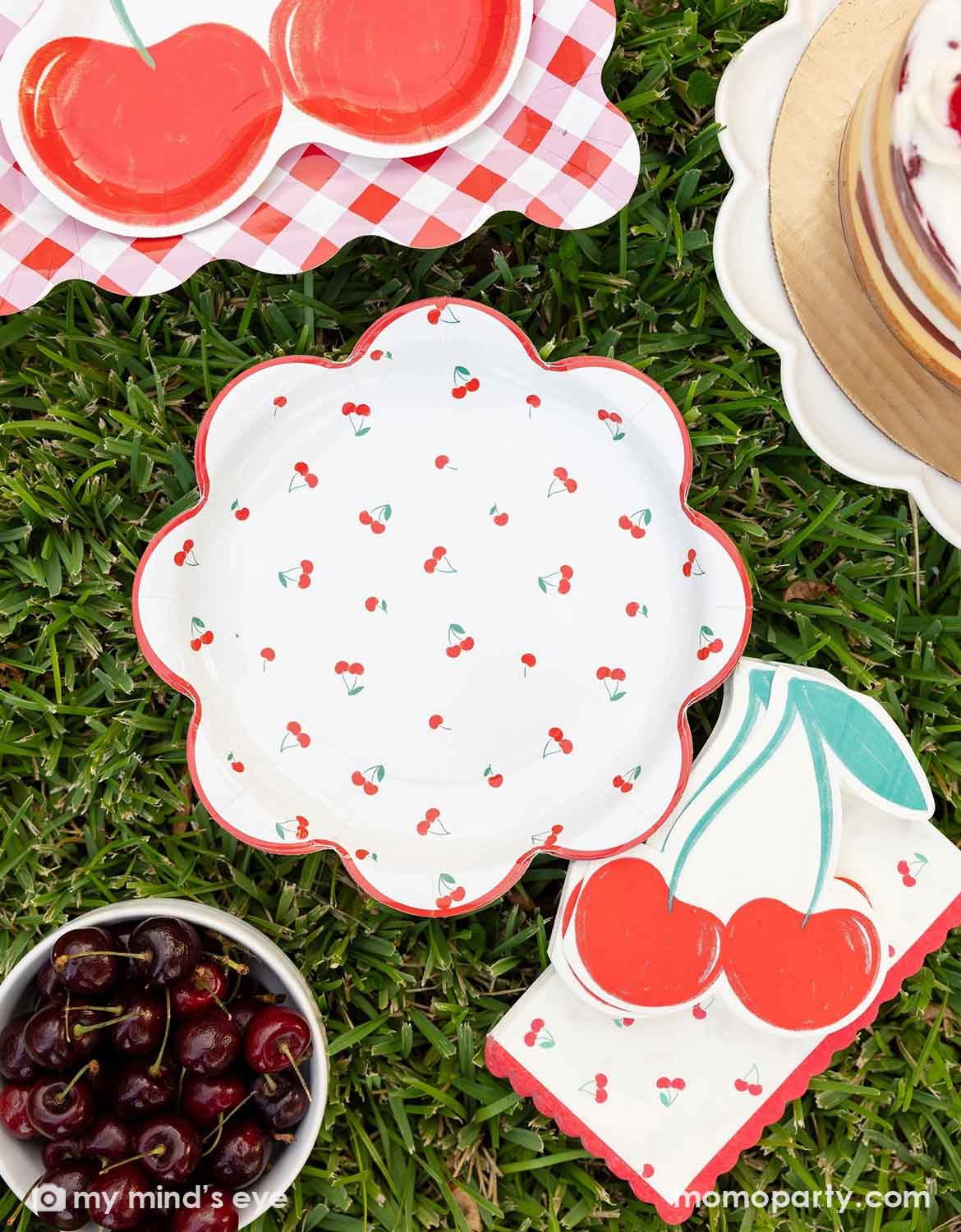 Momo Party's cherry-themed party plates and napkins by My Mind's Eye on grass in a picnic setting with a cake, and a small bowl of cherries around.
