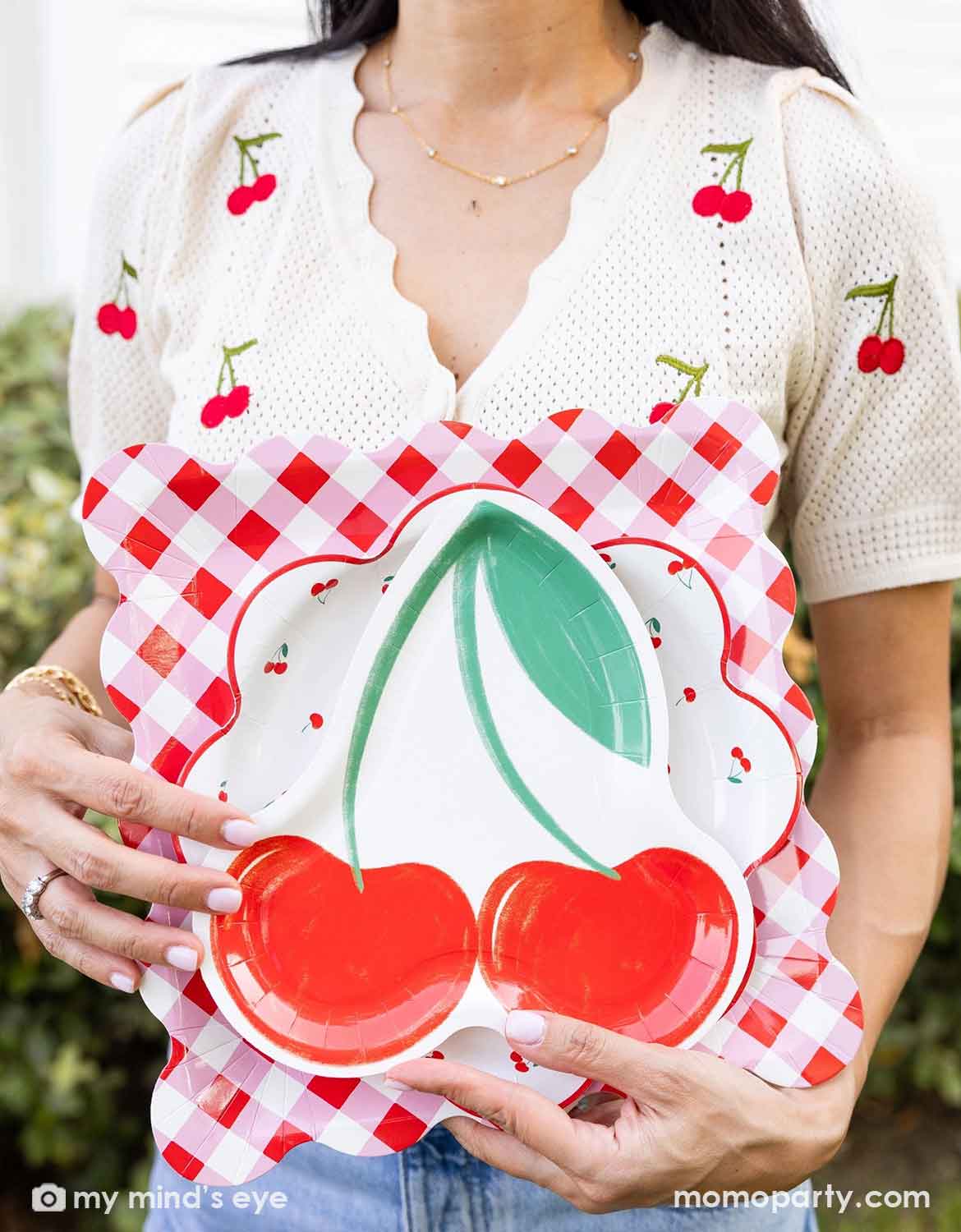 A lady in a shirt with cherry pattern on it  holding a Momo Party's cherry themed plates with cherry design and checkered pattern by Momo Party standing at an outdoor party venue.