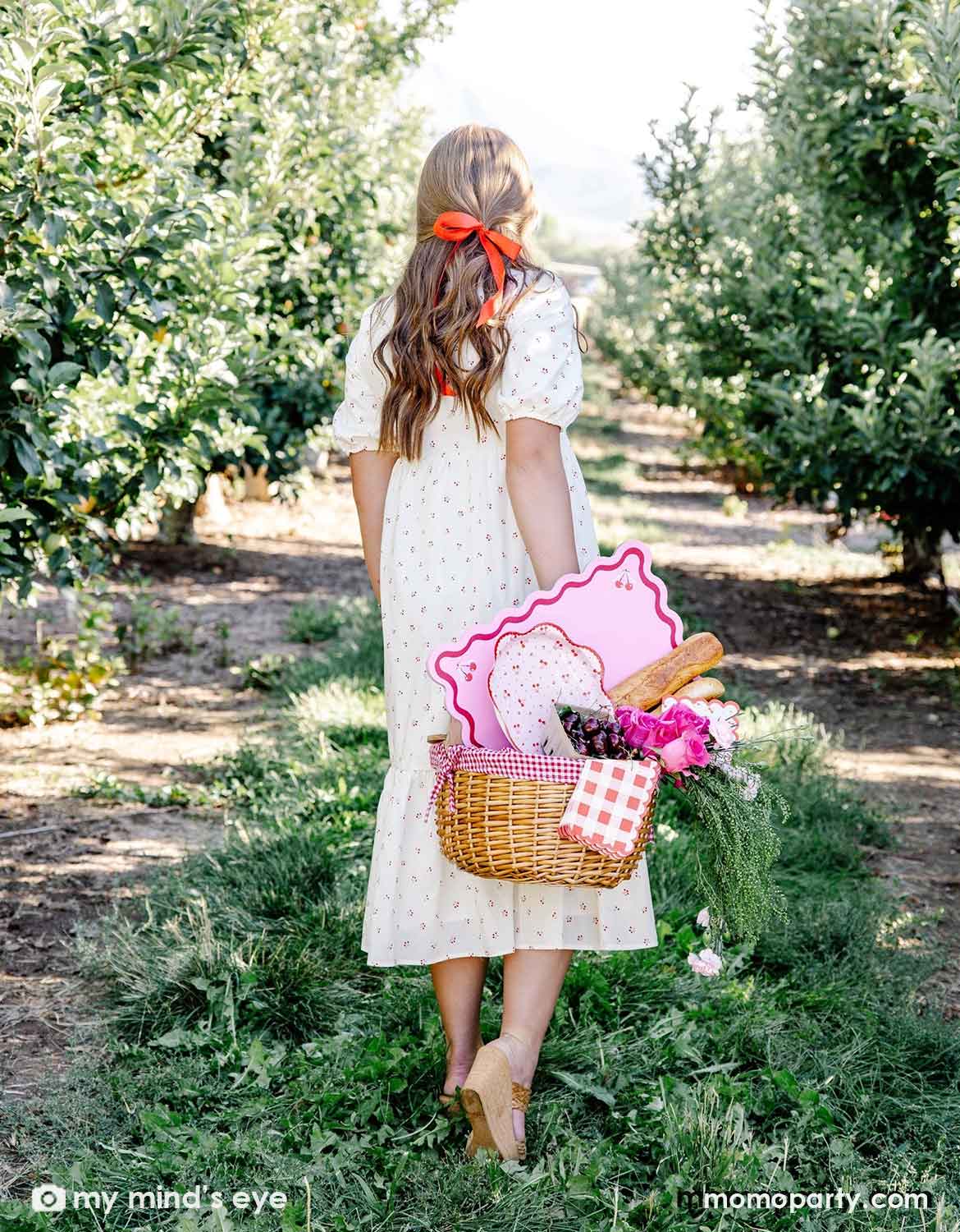 A lady in a white dress with a red cherry pattern holding a picnic basket filled with cherry themed party supplies including plates, napkins and placemats from Momo Party along with some bread and flowers walking through an orchard.
