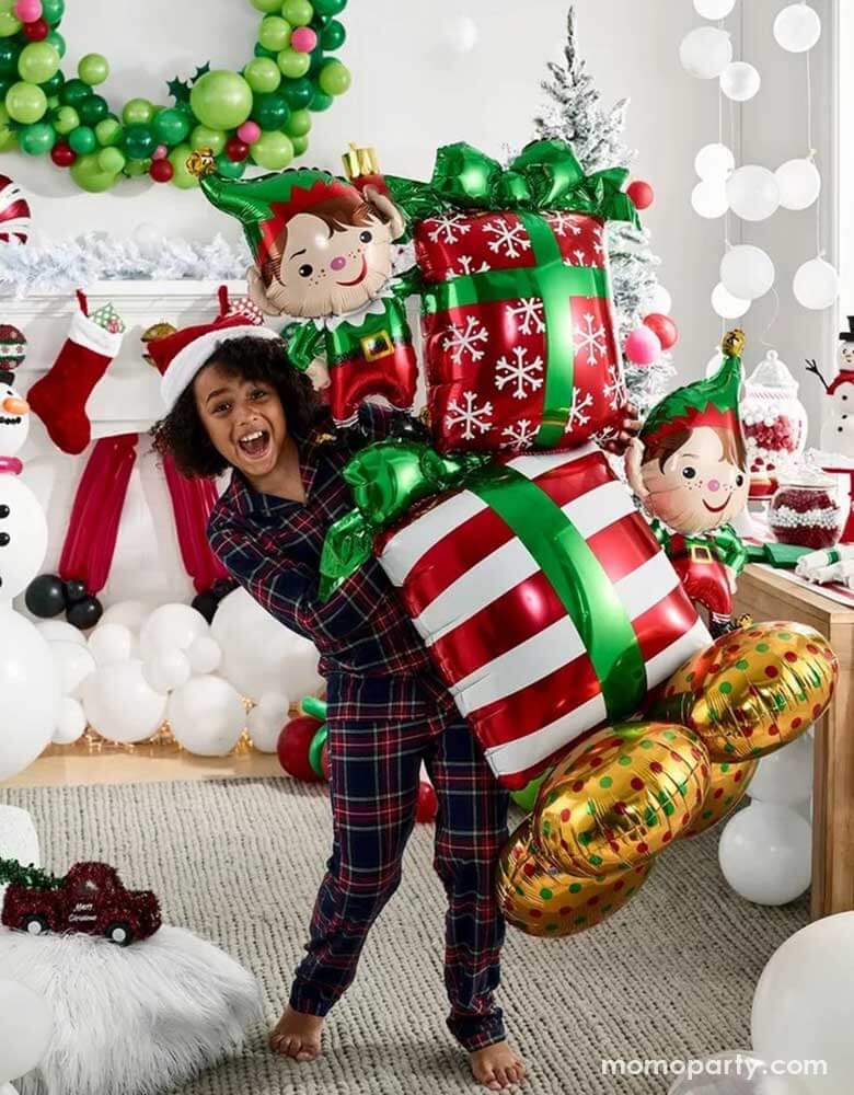 A girl in her Holiday pajama holding Momo Party's 36" x 53" Christmas Elves Airloonz balloon by Anagram Balloons. In the back it's a room decorated with Christmas balloons including a wreath, a snowman and some white balloon garland on the floor resembles snow. In the mantle there are stockings with gifts inside, makes it a perfect inspo for a fun Holiday gathering set up.
