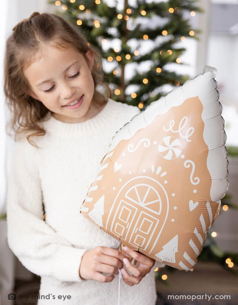 A girl in her cream white sweater holding Momo Party's 13.5" x 20" gingerbread house shaped foil balloon standing next to a big window. Behind her is a big Christmas tree lit up with small twinkling lights,