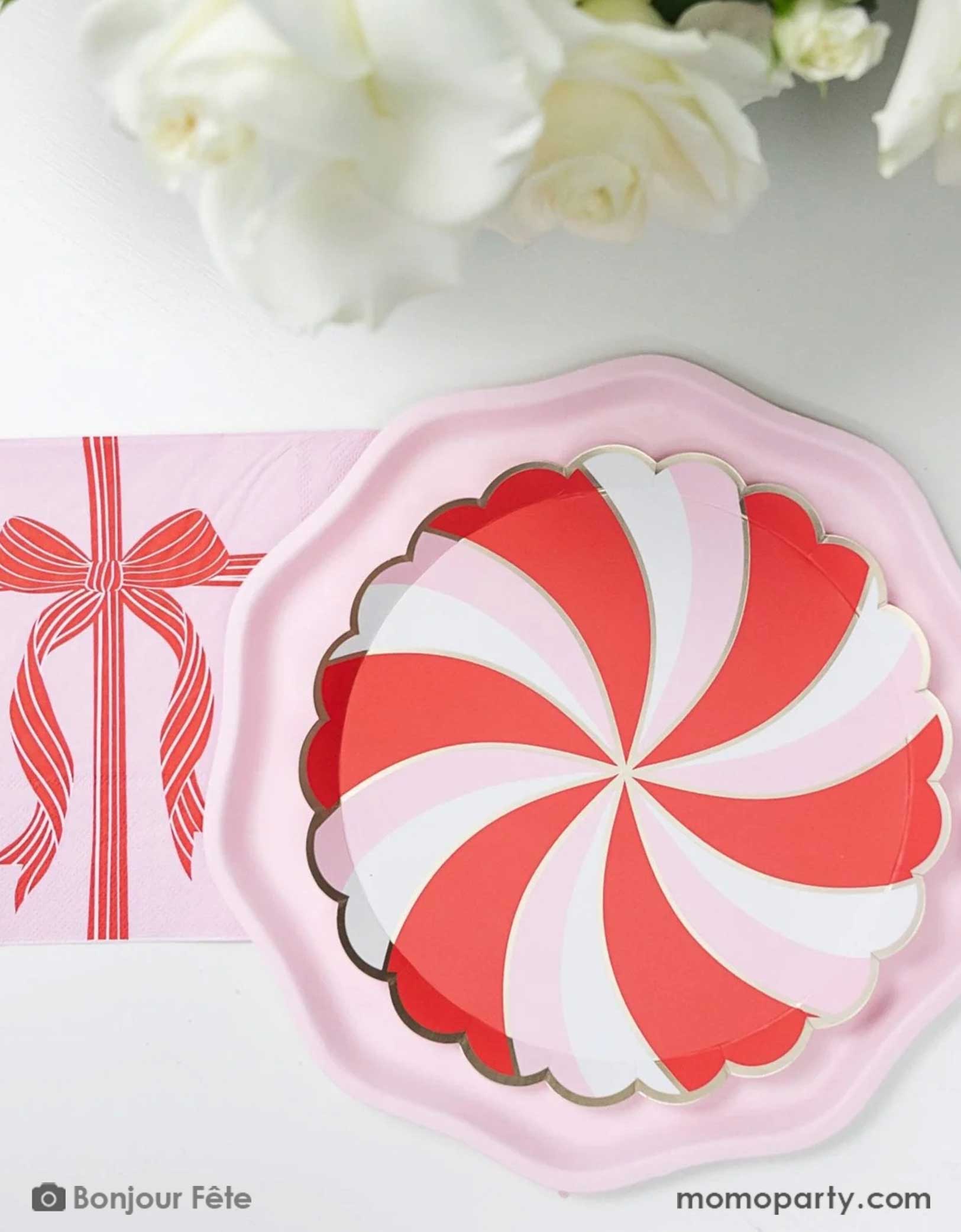 A Holiday party tablescape featuring Momo Party's peppermint side plate with red and white candy swirl design on a pink dinner plate, along with a pink large napkin with a red bow design on a white table with floral background.