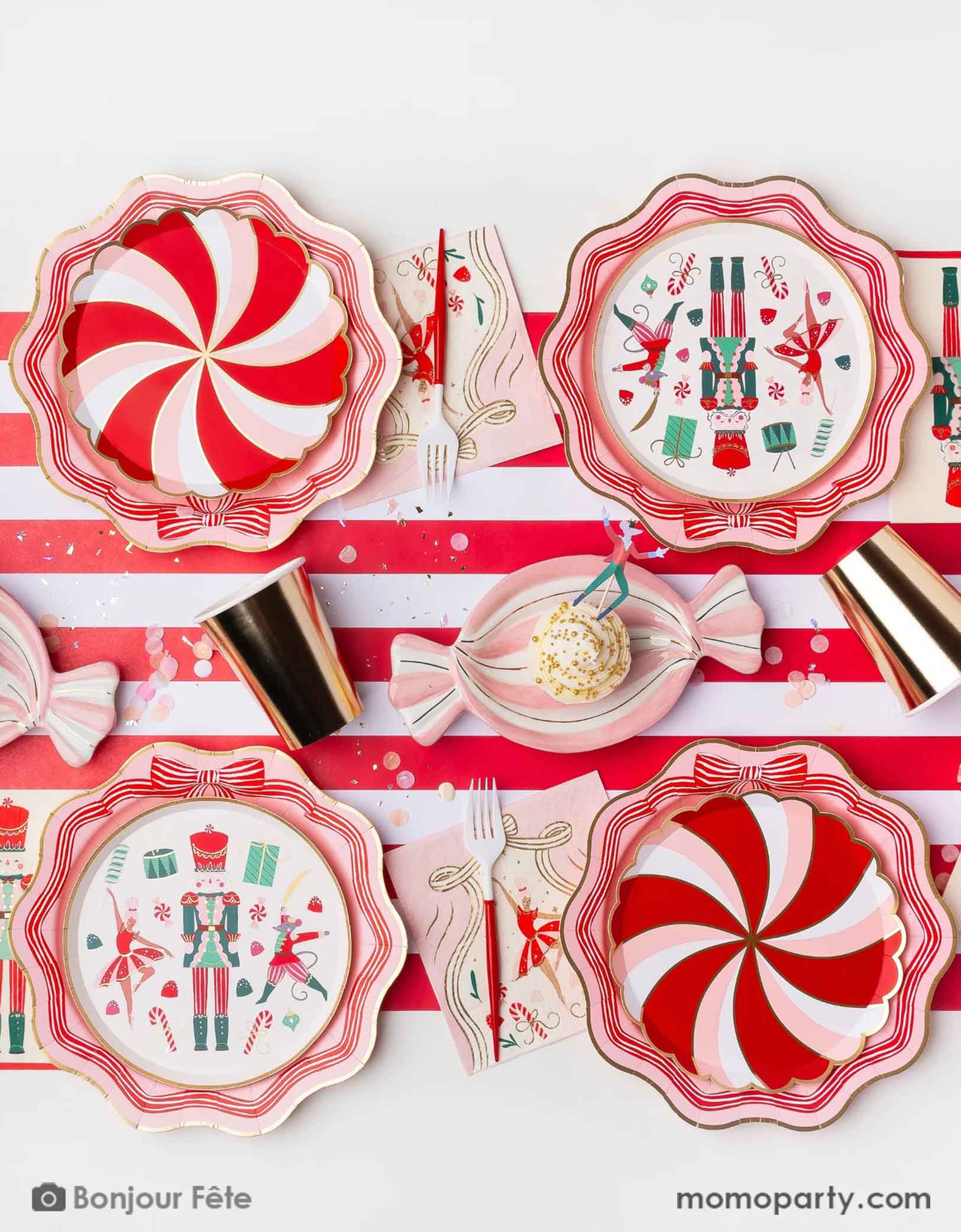 A festive Holiday candy themed party table setting featuring red and white peppermint plates, Nutcracker plates and decorative napkins and red and white utensils with candy-themed designs on a matching tablecloth from Momo Party.