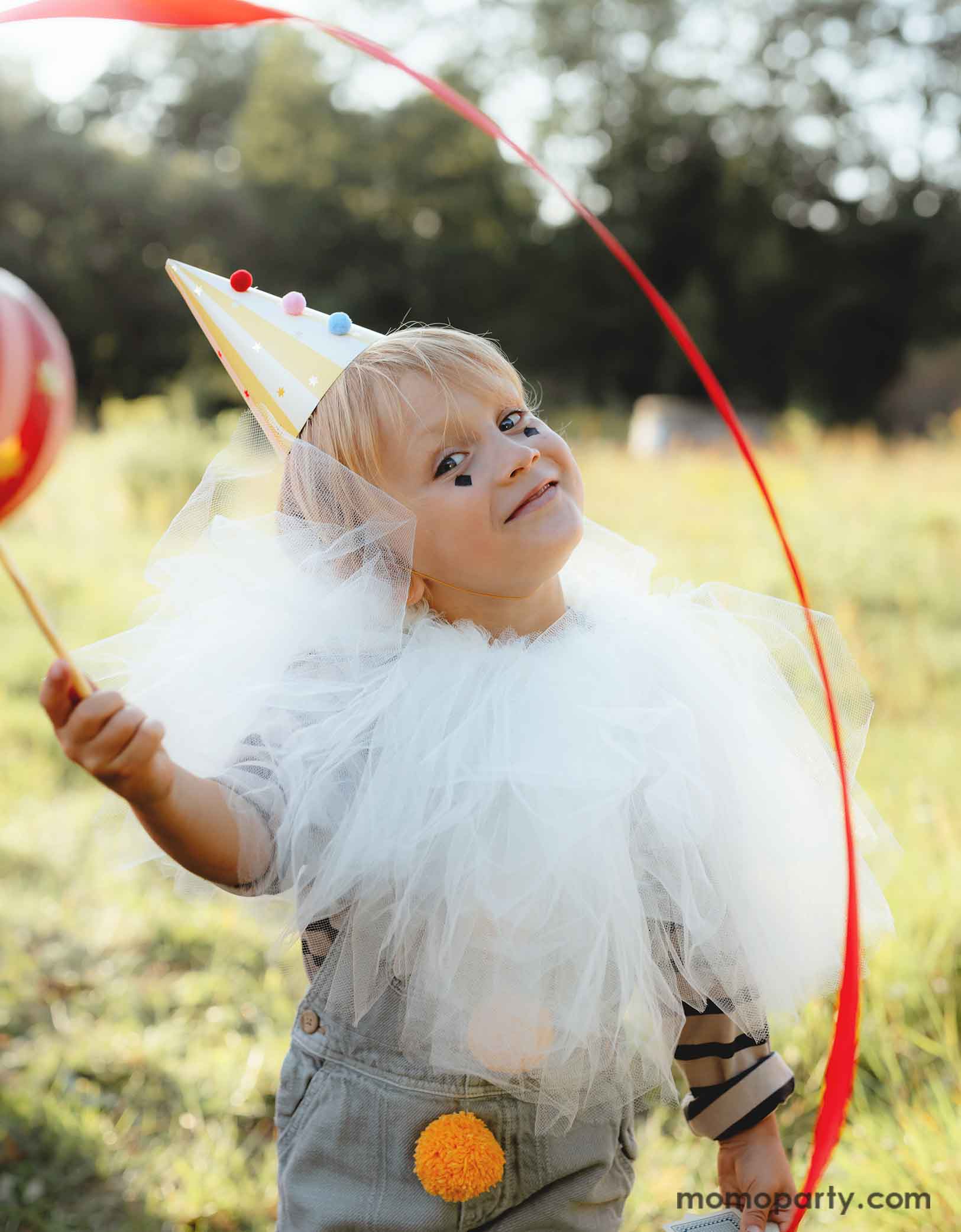 A little boy dressed in dreamy white tulle cover with vintage circus themed outfits wearing Momo Party's yellow striped Birthday Party Hat with Pompoms, in a festive carnival themed event. He's waving a red party streamer in the field and having fun. 