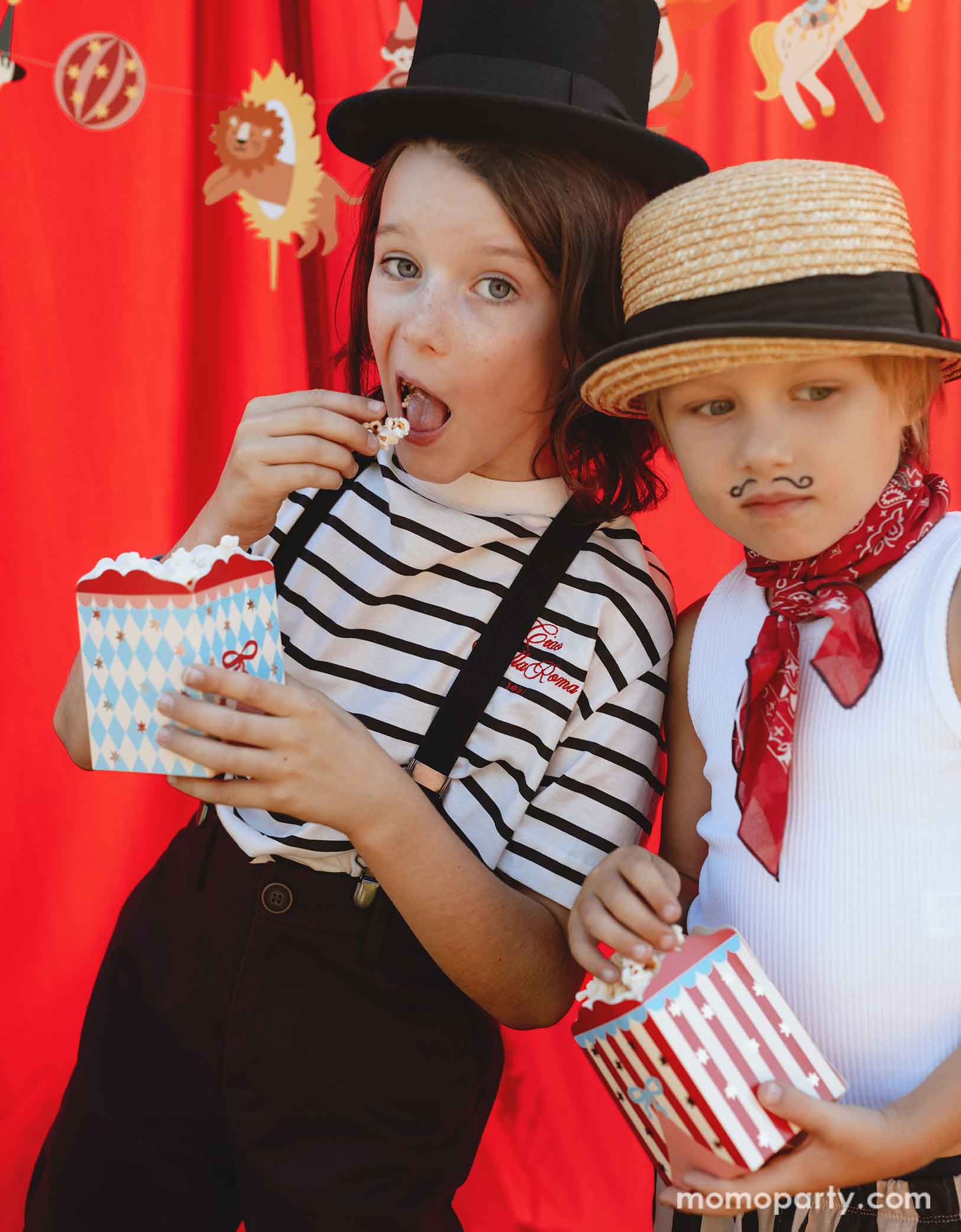 Two kids dressed in the vintage circus outfits and makeup holding Momo Party's Circus Striped Treat Boxes filled with popcorn in their hands in a festive carnival themed event. In their back is a red stage curtain as a party backdrop decorated with circus themed birthday banner with circus animal party pennants. 