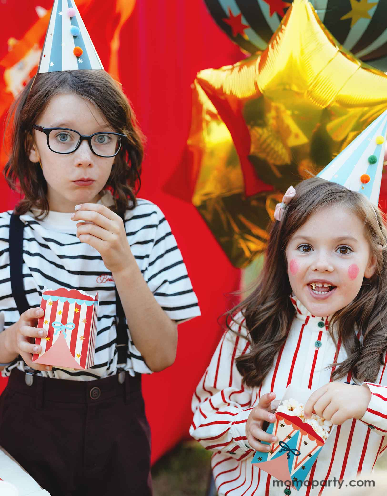 Two kids dressed in the vintage circus outfits and makeup holding Momo Party's Circus Striped Treat Boxes filled with popcorn in their hands, wearing party hats with colorful pom poms in a festive carnival themed event. In their back is a red stage curtain as a party backdrop decorated with circus themed birthday banner with circus animal party pennants and some festive large carnival themed foil balloons. 