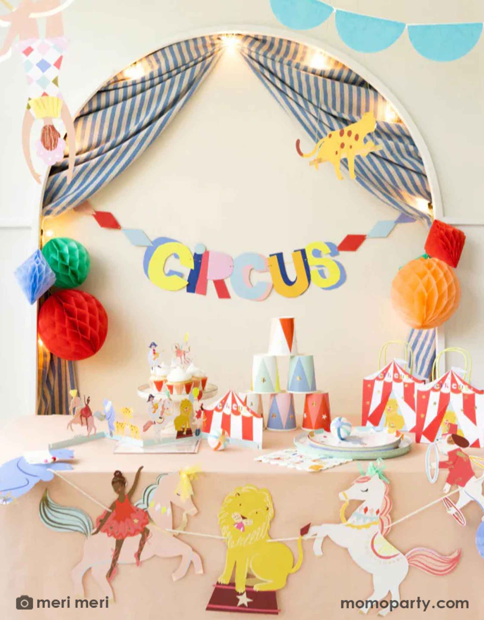 A festive circus themed party set up featuring Momo Party's circus themed party supplies including a table filled with circus themed party tablewares including plates, napkins, party cups, party bags and ferry's wheel cupcake stand. There's the circus party garland featuring circus animals with honeycomb decorations hung above the party table. 