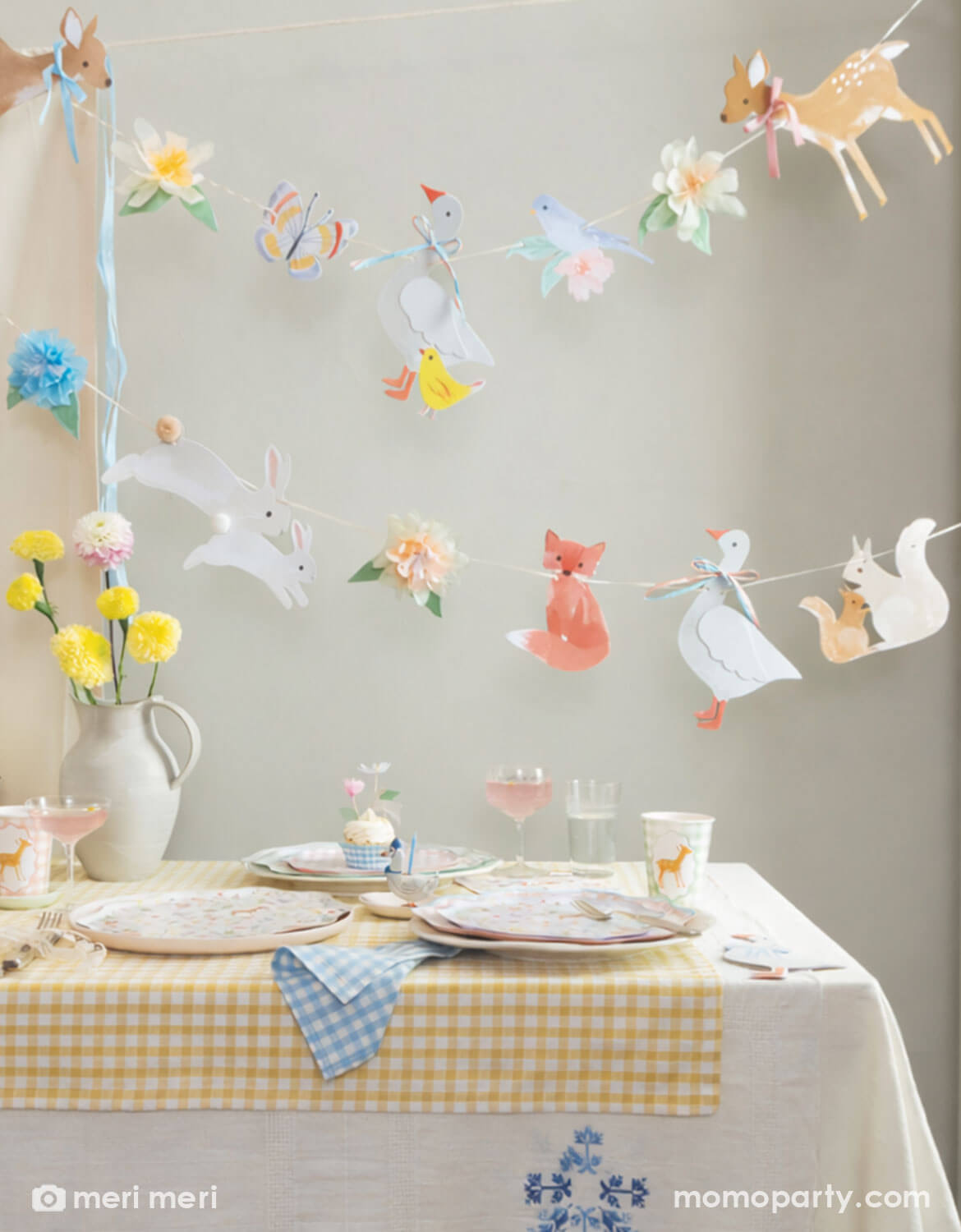 A rustic cottage themed table setting with little woodland animals of deer, goose, bunny, fox, chicks in tablewares and decorations from Momo Party - including party cups, matching plates and napkins and an adorable little animals party garland hung above the table covered with a soft gingham tablecloth. With spring florals in soft yellow and pink on the table, this table is perfect for a baby shower, an Easter celebration or young kid's storybook rustic cottage inspired birthdays.