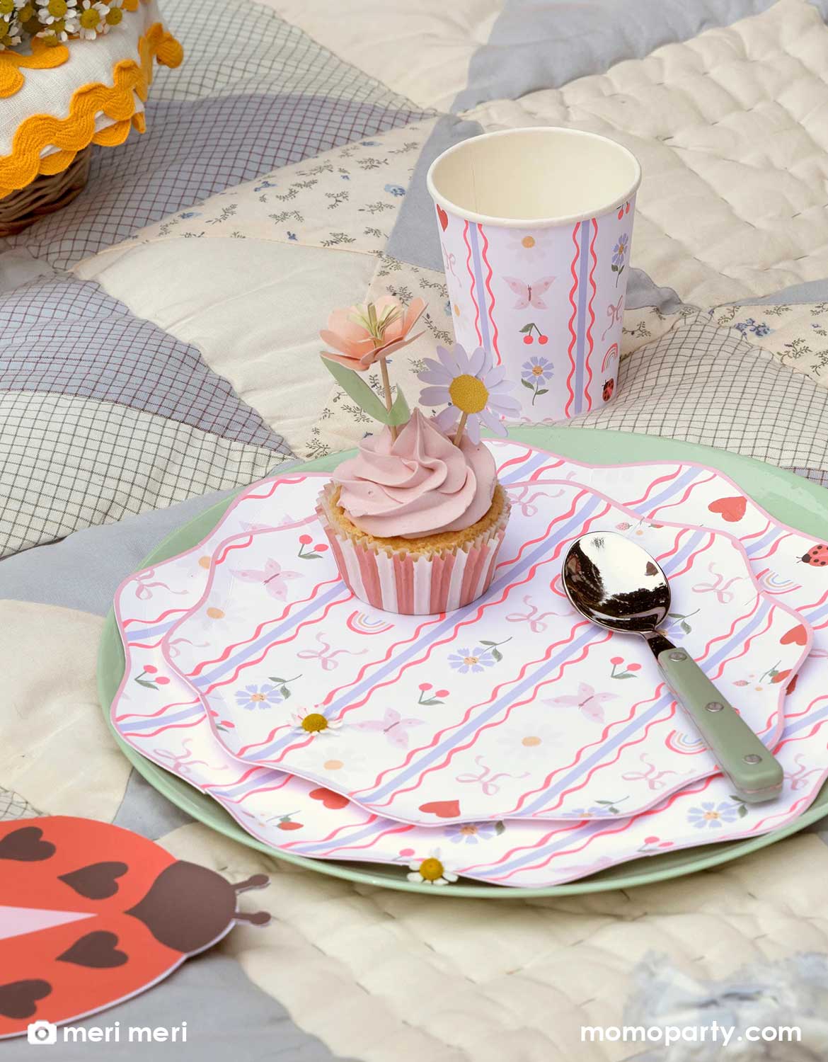 A rustic cottage inspired table setting featuring Momo Party's Little Garden themed tablewares including plates, cups and napkins featuring textile-inspired pattern of ladybugs, hearts, bows, flowers, cherries, butterflies and rainbows. There's a cupcake with pink frosting and a spoon, on a rustic themed quilt tablecloth.