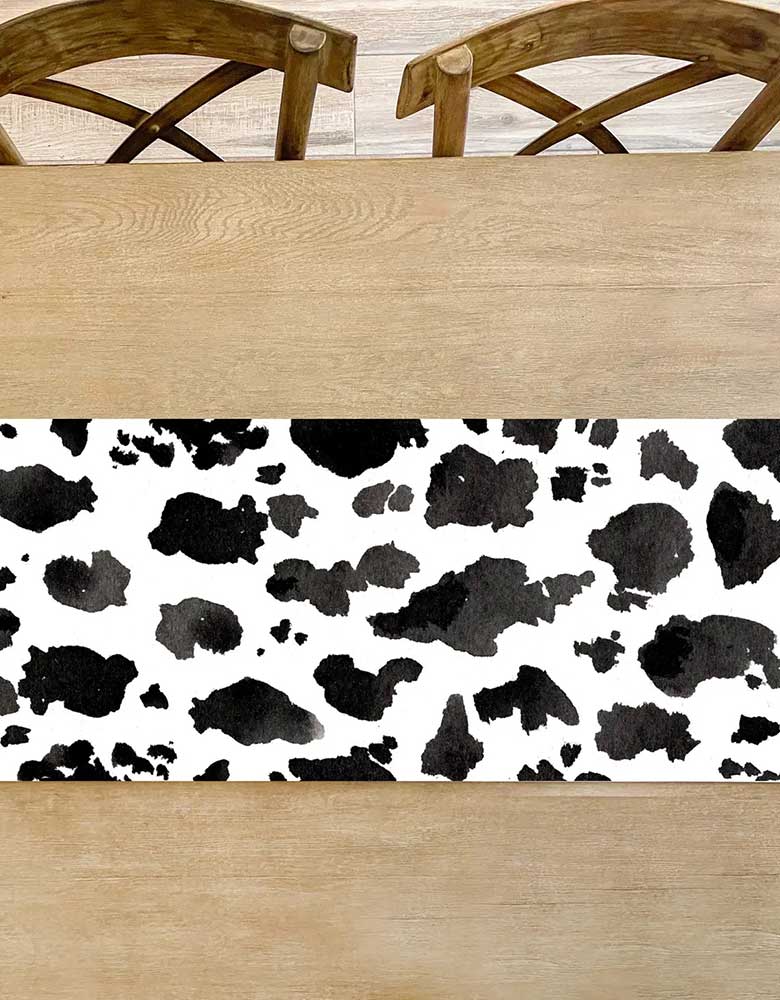 Momo Party's 16.5" x 96" cow print watercolor table runner by Cami Monet on a farm house style wooden table. This watercolor cow print table runner is the perfect addition to any western-themed celebration! With its eye-catching design, this table runner is guaranteed to bring a touch of rustic charm to your party decor. Perfect for cowgirl bachelorette parties, cowboy birthday parties, rodeo parties, farm birthday party themes and more.