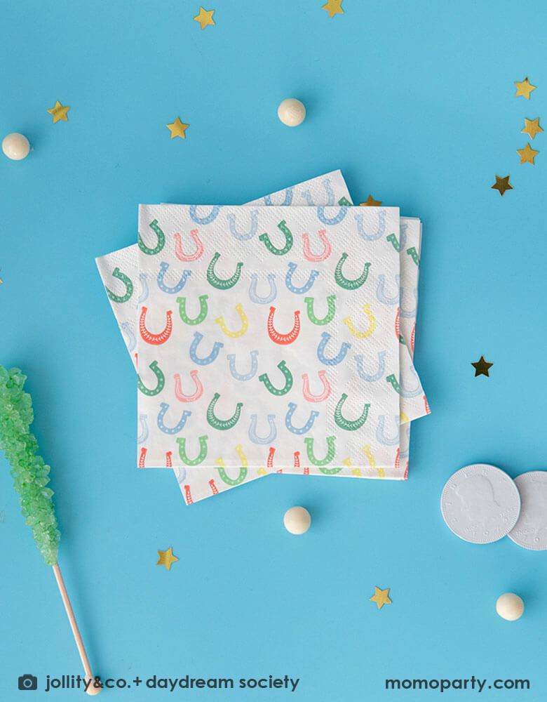 Momo Party's 5" x 5" horseshoe small napkins by Daydream Society on a bright blue table with gold star foil confetti and white gum balls surrounding them. With a green rocky ice lollipop and white chocolate coins next to the napkins. 