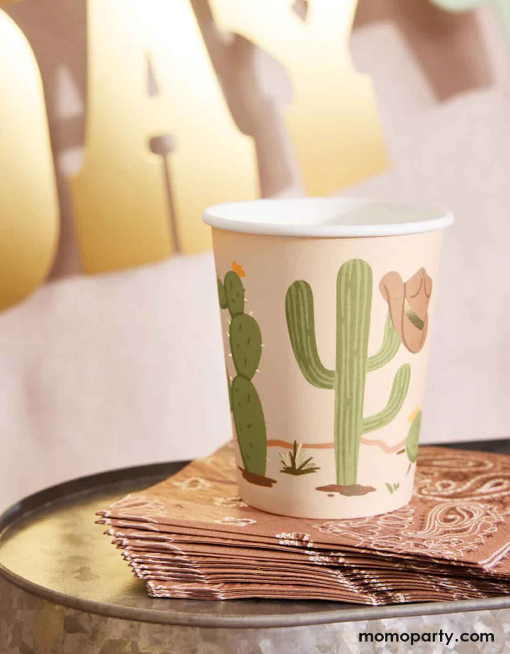 Momo Party's 9oz Wild Wild West Party Cup with Western cowboy and desert, cactus design sitting on a set of brown  small napkins with bandana pattern on a metal bucket. In the back is a gold foil Happy Birthday party banner hung on the wall.