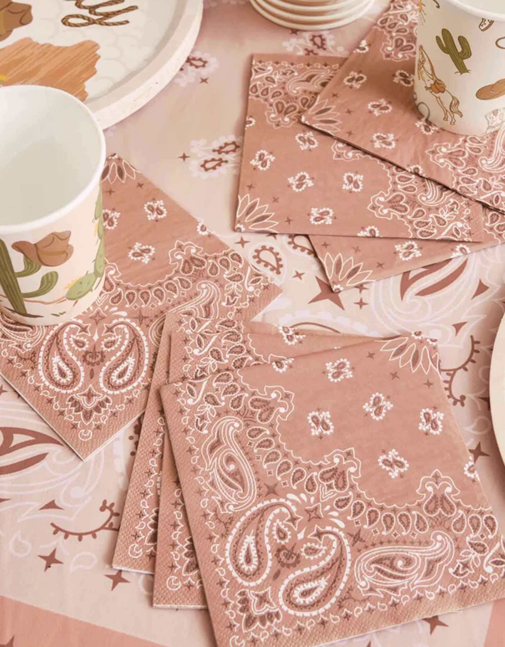An overhead shot of Momo Party's Western cowboy rodeo themed party tablewares featuring 5" bandana patterned napkins and Western themed party cups on a table covered with a beige tablecloth with bandana design.