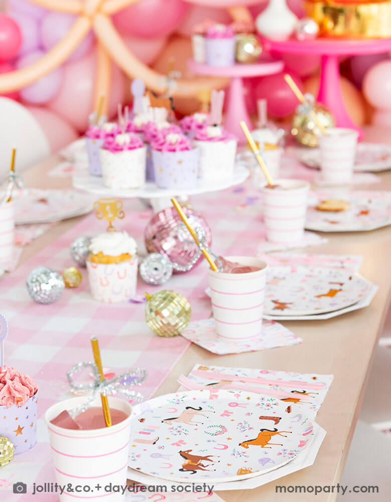 A close up of a girly cowgirl rodeo pony themed party table featuring Momo Party's cowgirl pony themed party plates, napkins, horseshoe napkins, pink striped party cups and a pink gingham table runner. Around the tableware there are disco ball decorations and a cake stand with cupcakes with pink icing. Behind the table there is a balloon wall with pink balloons, making this an adorable inspiration for girl's First Rodeo first birthday celebration or an adorable cowgirl theme party for pony-loving girls!