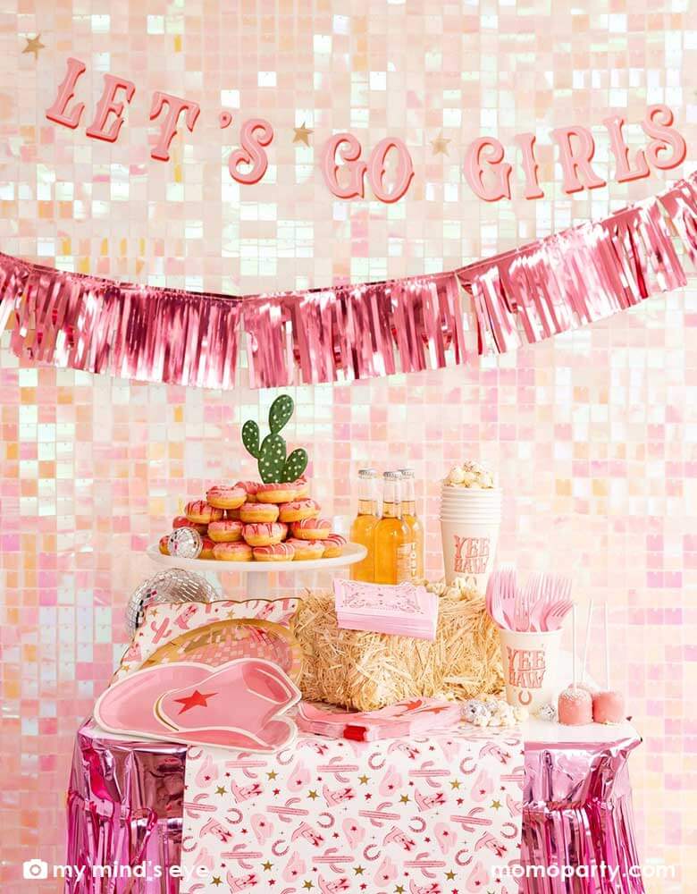 A cowgirl Western themed party table features pink cowgirl themed party supplies from Momo Party including pink cowgirl boot shaped napkins, cowgirl hat shaped plates, pink disco ball shaped plates, pink bandana small napkins, yeehaw party cups and cowgirl pattern paper plates and table runner by My Mind's Eye. In the back there's an iridescent backdrop adorned with Let's Go Girls pink foil fringe banners, along with a pink donut tower cake, making this a great inspo for a disco cowgirl party.