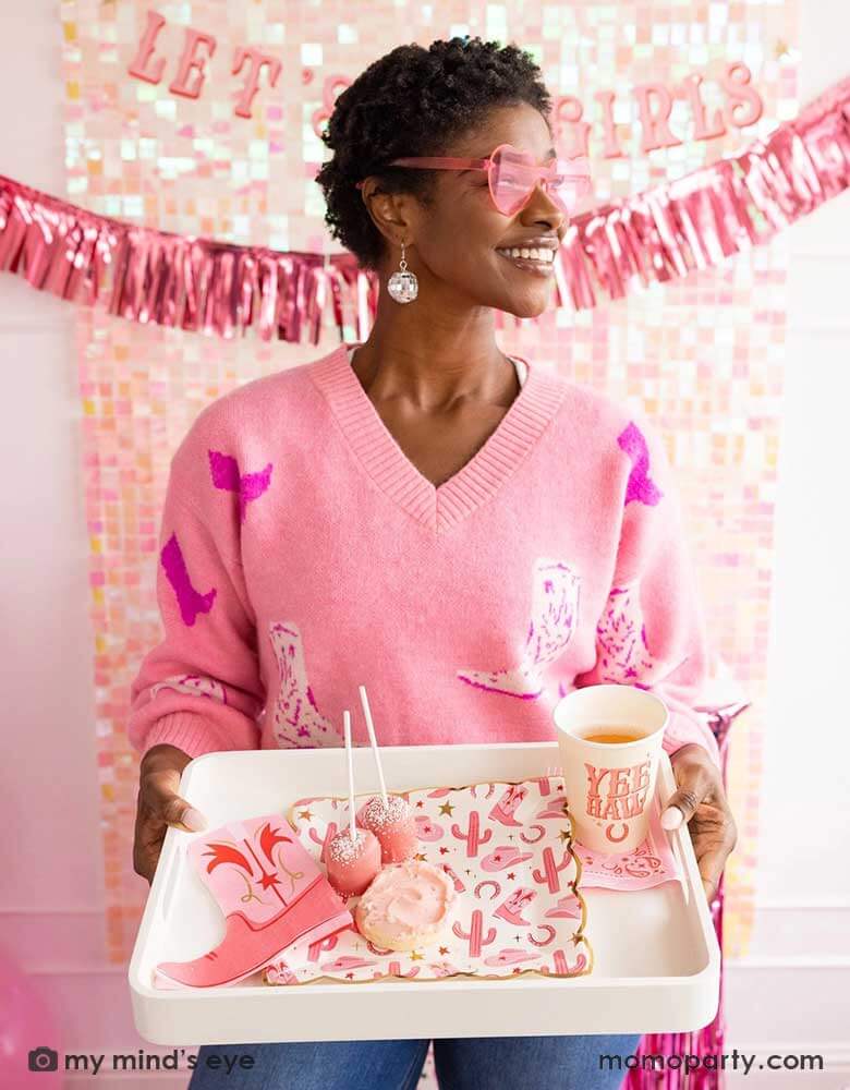 A young African American lady in a pink sweater with cowgirl boot pattern on it with disco ball ear rings and pink heart shaped sunglasses on her holding a tray with cowgirl party supplies from Momo Party including cowgirl pattern plates, pink cowgirl boot shaped napkins, yeehaw party cups and some pink treats. In her back there's an iridescent curtain wall adorned with My Mind's Eye's Let's Go Girls party banner with pink foil fringe banner, making this a perfect inspo for disco cowgirl bachelorette party.