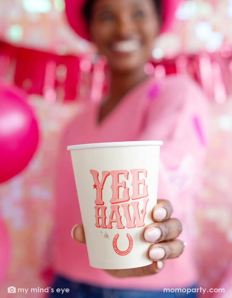 A young lady standing in front of a wall decorated with pink party banners and balloons, he's wearing a pink sweater with a pink cowgirl hat holding the 12 oz YEEHAW cowgirl party cup from Momo Party, enjoying herself in this disco cowgirl themed party.