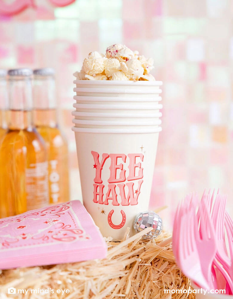 A cowgirl rodeo Western themed party table features pink cowgirl themed party supplies from Momo Party including 12oz pink YEEHAW party cups with popcorn in them, pink bandana small napkins and some soda bottles on a stack of hays, making this a perfect inspo for kid's cowgirl themed birthday party or a disco cowgirl bachelorette party celebration!