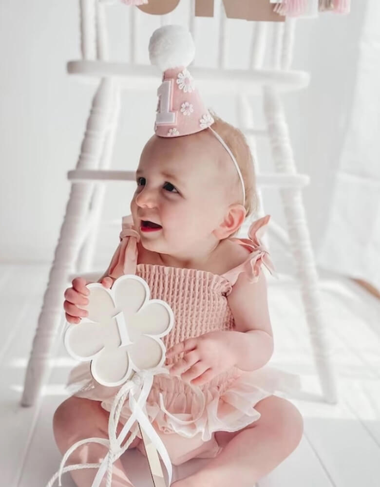 A little baby girl in adorable pink onesie wearing Momo Party's Daisy Applique felt birthday hat on her head. In her hands, she is holding a flower shaped pennant with number 1 on it. She's sitting in front of a vintage inspired white highchair celebrating her first birthday.