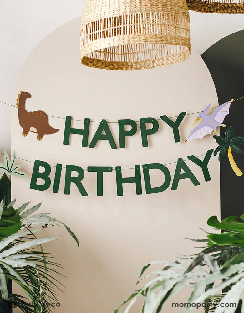 A kid's dinosaur themed party backdrop wall decorated with Momo Party's dinosaur happy birthday banner by Party Deco. This green happy birthday banner featuring dinosaur and palm tree pennants is perfect to set a scene for kid's dinosaur, Jurassic themed birthday party.