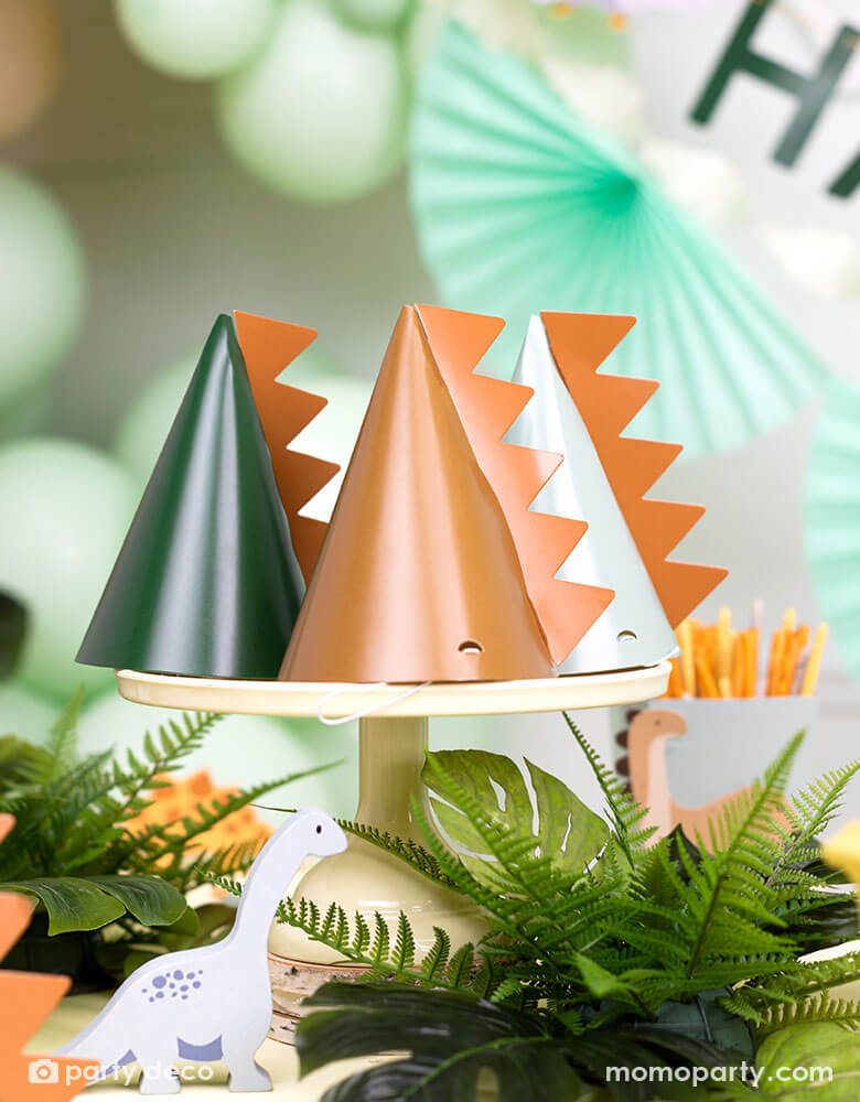 A festive kid's dinosaur themed party table features Momo Party's 5.5" Dinosaur Party Hats by Party Deco. Comes in a set of 6 party hats in 3 different colors of green, mint, and brown, these dinosaur party hats are with a dinosaur spike on them. The table is decorated with jungle themed plants with mint, brown and green colored decorations on the wall including some party balloons and a festive bunting garland.