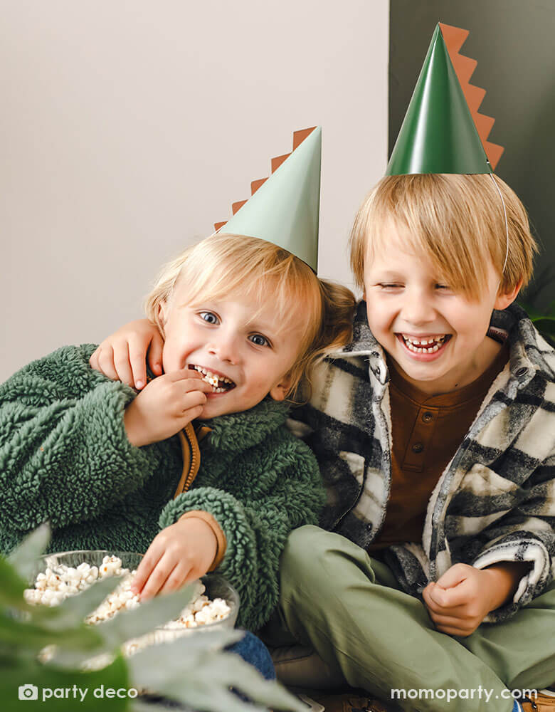 Two school aged boys in green themed outfit wear Momo Party's 5.5" Dinosaur Party Hats with a dinosaur spike on them by Party Deco. They're enjoying the popcorn from the party, laughing and having a good time.
