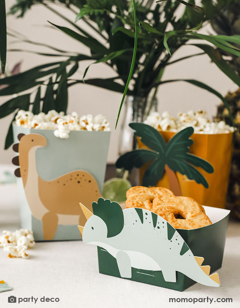 A kid's dinosaur party table features Momo Party's dinosaur snack boxes filled with cookies and popcorn, around the table are some tropical plants, creating a Jurassic pre-historic vibe for this dinosaur adventure.