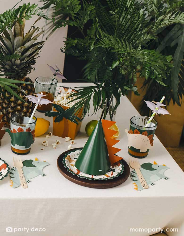 A kid's dinosaur themed party table features Momo Party's dinosaur themed party supplies including Triceratops shaped napkins, dinosaur snack boxes filled with popcorn, dinosaur cup sleeves topped with Pterodactyl paper straws and dinosaur party hats with dinosaur spikes. The table is decorated with tropical plants and pineapples, creating a Jurassic vibe for this kid's dinosaur party.