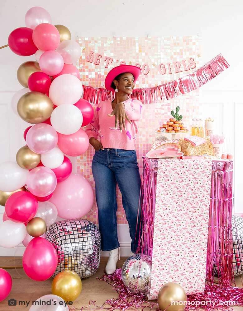A young African American lady in the Western inspired outfits and a pink cowgirl hat in a disco cowgirl bachelorette party decorated with cowgirl themed party supplies, decorations and balloons from Momo Party. She's showing her engagement ring next to a table adorned with pink fringe and cowgirl party supplies including plates, napkins and a paper table runner. There's a donut cake, beverages and various treats on the table. Behind them is a iridescent wall with "Let's Go Girls" banner from Momo Party.