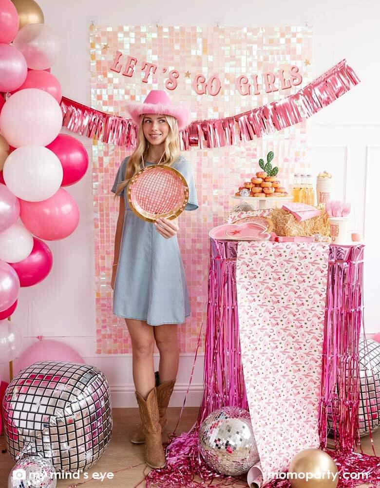 A young Caucasian lady in the Western inspired outfits and a pink cowgirl hat in a disco cowgirl bachelorette party decorated with cowgirl themed party supplies, decorations and balloons from Momo Party. She's holding the pink disco ball shaped plate by a table decorated with cowgirl party supplies including plates, napkins and a paper table runner. There's a donut cake, beverages and various treats on the table. Behind them is a iridescent wall adorned with "Let's Go Girls" pink foil fringe party banner.