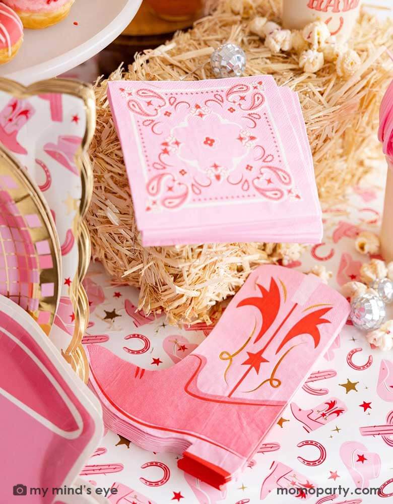 A cowgirl rodeo Western themed party table features pink cowgirl themed party supplies from Momo Party by My Mind's Eye including pink cowgirl boot shaped napkins, pink bandana small napkins, cowgirl hat shaped plates, yeehaw party cups and cowgirl pattern paper table runner, along with hay stacks and small disco ball decorations, this makes a perfect inspo for kid's cowgirl themed birthday party or a bachelorette party celebration!