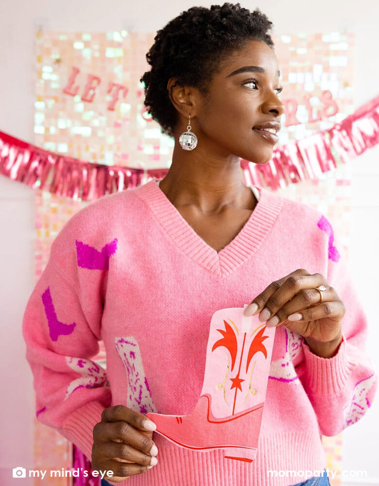 A young African American lady in a pink sweater with cowgirl boot pattern on it with disco ball ear rings on her holding Momo Party's pink cowgirl boot napkins. In her back there's an iridescent curtain wall adorned with My Mind's Eye's Let's Go Girls party banner with pink foil fringe banner, making this a perfect inspo for disco cowgirl bachelorette party celebration.
