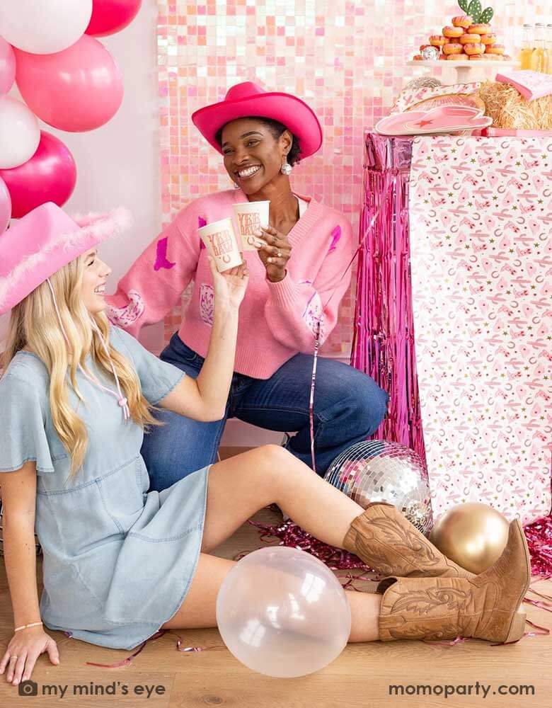 Two ladies in their Western rodeo cowgirl inspired outfit in a disco cowgirl themed bachelorette party decorated with cowgirl themed party supplies, decorations and balloons from Momo Party. They're raising their 12oz YEEHAW party cups by My Mind's Eye enjoying themselves in this festive celebration with donut cakes, beverages and various treats on the table decorated with cowgirl pattern table runner and pink foil fringe next to them. 